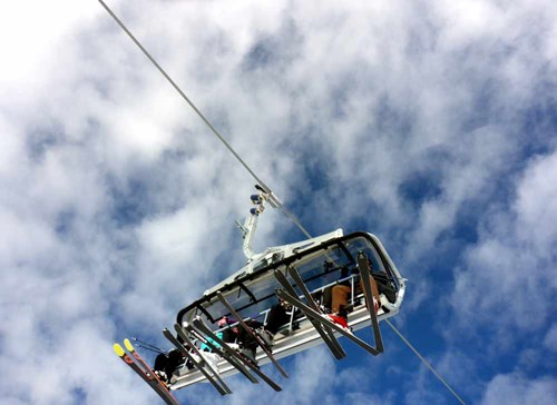 A group of skiers in a ski lift dangling their feet of the end, in the background is a cloudy sky