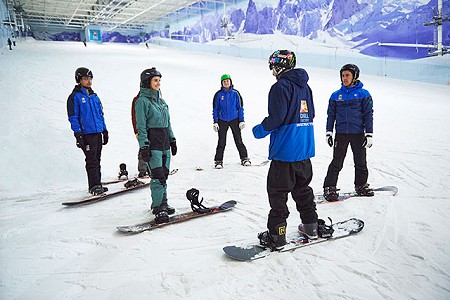 a group of adults on a snowboarding lesson stood around listening to their instructor