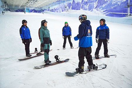Group of adult snowboarders on the main slope at Chill Factor<sup>e</sup> stood listening to their instructor.