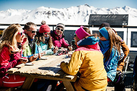 Group of skiers enjoying a drink around a table with mountains in the background