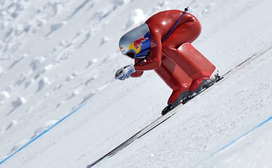 A skier in streamline red clothing and a red bull helmet, crouched down going down the slope