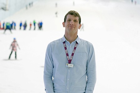 Man in blue shirt with purple lanyard, in the background people are skiing down a slope in the snow.