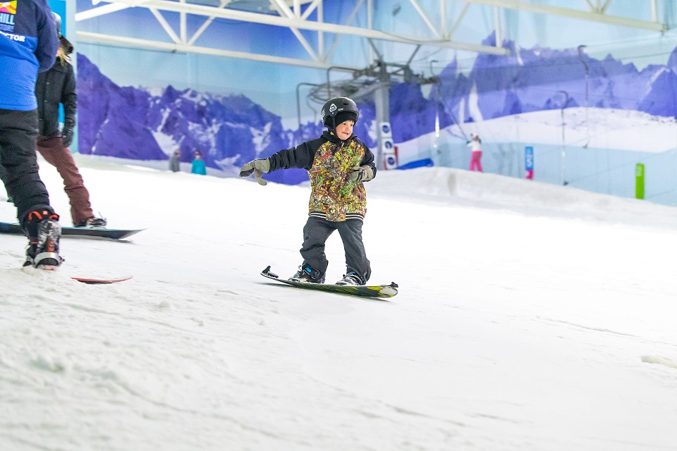 young kid in a multicolour coat and black helmet snowboarding on the slope