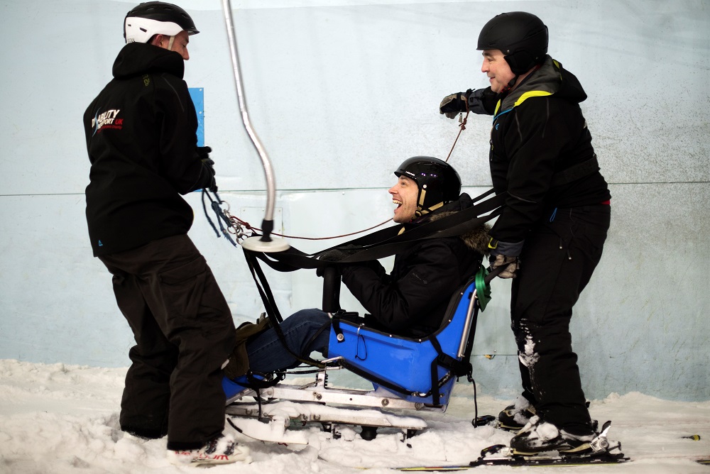 two people stood in black helping a disabled man in a sit-ski, the man is smiling at them