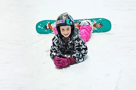 Child lying on snow with snowboard attached, smiling to camera