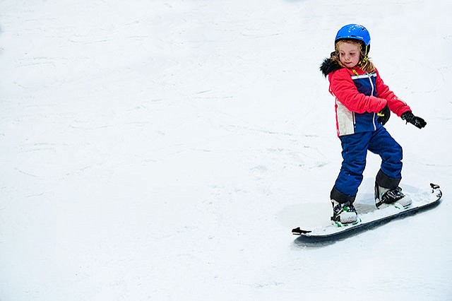 young girl snowboarding down the slope in her blue pants, red jacket, and blue helmet