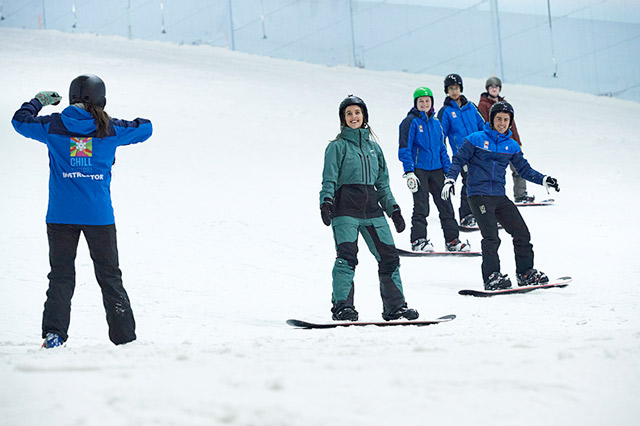 a group of adults on a group snowboarding lesson on the main slope stood looking at the instructor
