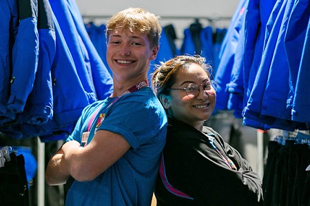 Man and woman stood back to back, behind them are lots of blue jackets on hangers.