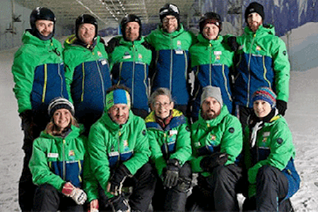 Eleven people in green coats and ski equipment posing for a group photo while on ski slope