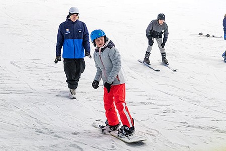 Young person sliding down the slope on a snowboard with instructor walking behind them