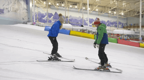 A man and woman on the indoor ski slope at chill factore, they are facing eachother and wearing hats