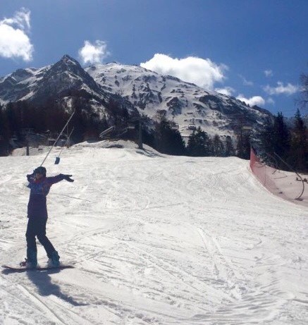 A skier stood in the snow on a slope infront of a large mountain with their arms in the air