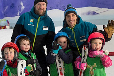 Two adults and four children stood together in the snow, the children are holding skis and smiling.