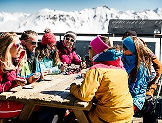 Group of people wearing ski clothing sat outside, around a table. Snow covered mountains in the background