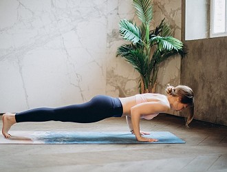 A lady in the plank position on a yoga mat, wearing black leggings and a pink sports bra