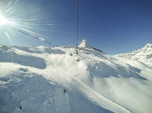 A range of snowy mountains with a ski lift, there is a clear blue sky and the sun in the corner