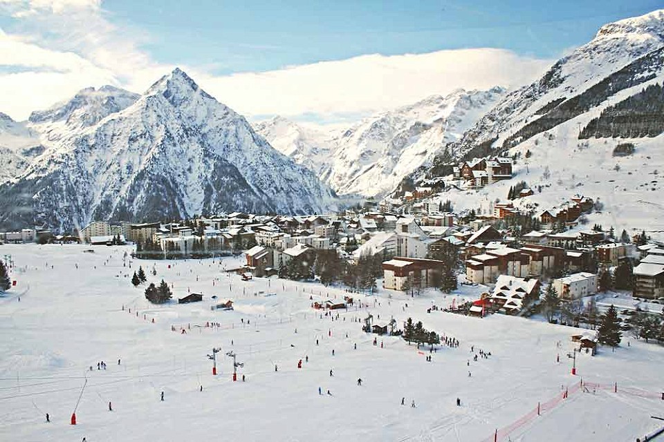 View of a ski village, in the foreground is a group of skiers, there are mountains in the background