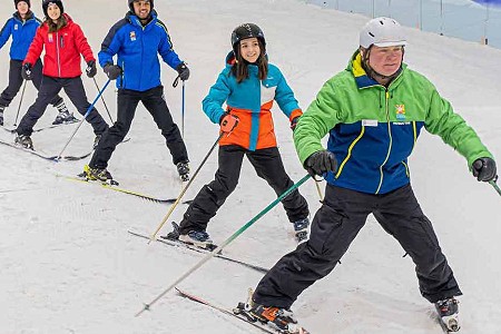 Five people skiing in a diagonal line on the snow, they are all wearing black trousers.