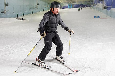 Man skiing in the snow, he is wearing a black jacket and trousers. He is on an indoor ski slope.