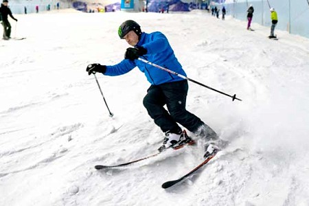A man in a blue jacket and black trousers, skiing down a slope and flicking up snow as they turn