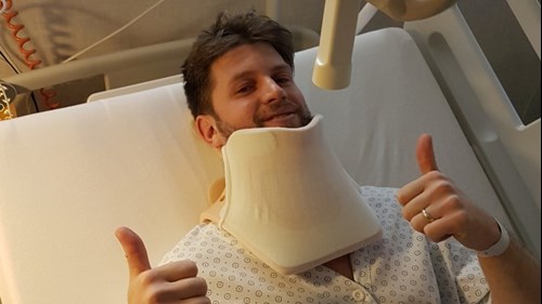 A man in a hospital bed wearing a neck brace, smiling at the camera with their thumbs up